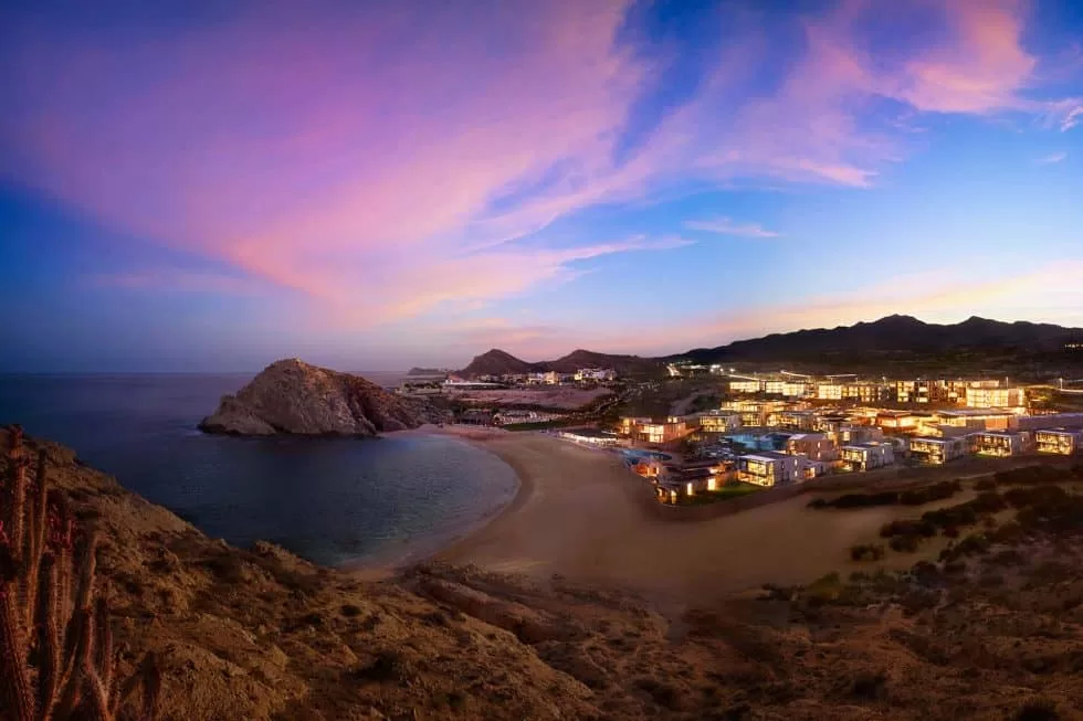 En un hermoso atardecer en Baja California, se vislumbra un horizonte con una playa de aguas cristalinas, el sol se refleja en el océano, mientras una lujosa villa en la costa se alza majestuosamente